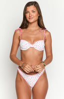 Girl is shown from above wearing a white bikini top with a small red floral pattern and red gingham trim along the underwired cups and on the adjustable tie shoulder straps. The matching bikini bottoms with gingham trim are also visible.