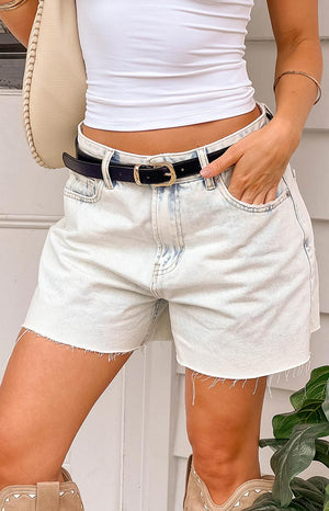 Girl wearing light wash denim shorts with a white tank top, black and gold belt, beige cowboy boots and holding a beige handbag