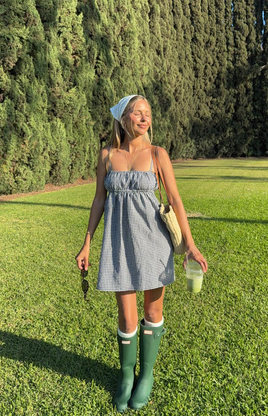 A woman in a black and white gingham mini dress with a ruched bust and spaghetti straps, accessorised with a white bandana, white socks, and dark green rain boots.