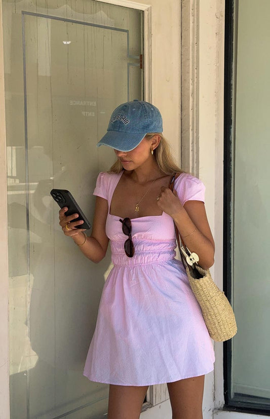 A woman in a light pink and white striped mini dress with cap sleeves and a square neckline, and a blue denim baseball cap.