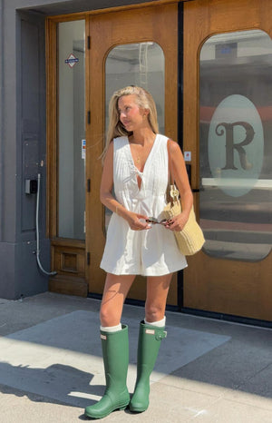 A woman stands outdoors in a sleeveless white playsuit featuring a V-neck with a tie at the bust and vertical pintuck detailing. She is wearing tall, matte green rain boots over white socks that peek out at the top.