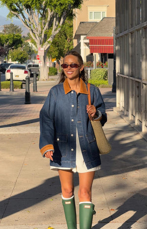 A woman walks on a sidewalk wearing an oversized, mid-wash denim jacket with a contrasting tan corduroy collar and corduroy trim on the cuffs and pocket flaps. A white outfit is visible underneath, paired with tall green rain boots and white socks.