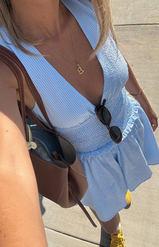 A close-up, high-angle view of a woman in a light blue and white pinstripe mini dress. The dress has a deep V-neckline and a wide, shirred elastic waistband.