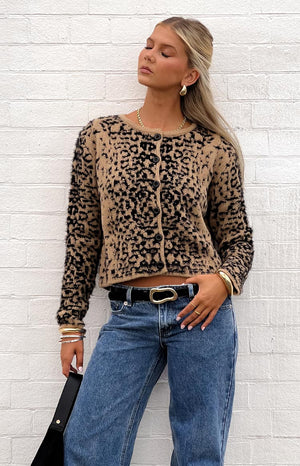 Woman wearing a textured tan and black leopard print button-up cardigan, blue jeans, a black belt with a gold buckle, and gold jewelry.