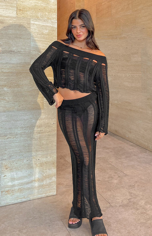Brunette model is posed in front of a brushed tiled wall. She is wearing a matching set, featuring a black, ladder knit, midi skirt. 