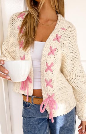 A close-up shows a person wearing a cream-colored chunky knit cardigan with pink cross-stitch accents and tie closures. They are also wearing a white top, light blue jeans with a brown belt, and a gold necklace while holding a white bowl.
