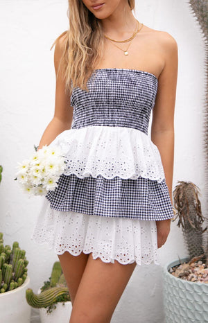 Model wearing a strapless mini dress with a navy and white gingham bodice and alternating white eyelet and gingham ruffled tiers on the skirt, holding white flowers.