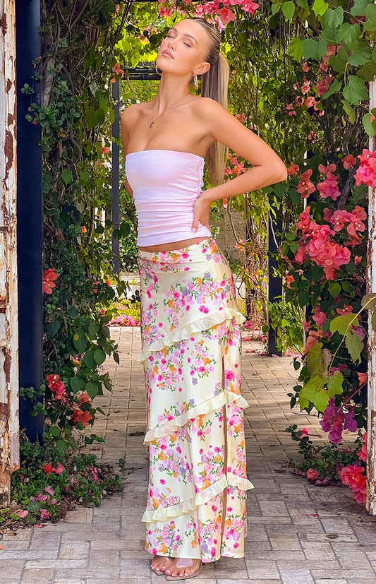 Blonde girl wearing a yellow floral maxi skirt and pink strapless top standing in a garden entrance.