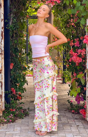 Blonde girl wearing a yellow floral maxi skirt and pink strapless top standing in a garden entrance.