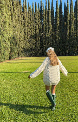Girl showing the back of an oversized cream cable-knit sweater dress. The image highlights the cozy, thick knit pattern and long, roomy sleeves.