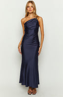 Our model wearing a floor length maxi dress in a dark navy blue satin fabric. The dress features an asymmetrical neckline with a draped cowl like bodice and thin spaghetti straps that crisscross over the back. The skirt is fitted through the hips and flows down to the floor.