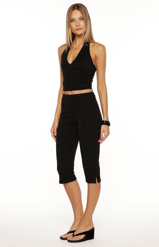 A close-up of our model wearing a pair of fitted high-waisted black capri pants. This bottoms have a smooth finish and a straight cut ending just below the knee with a small slit detail at the outer hem of the leg opening.
