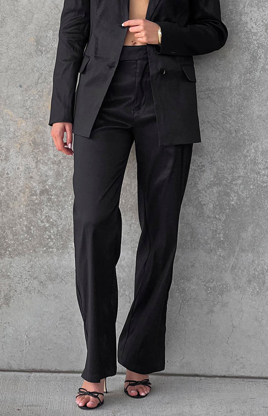 Model wearing black suit pants and blazer with black heels in front of a wall.